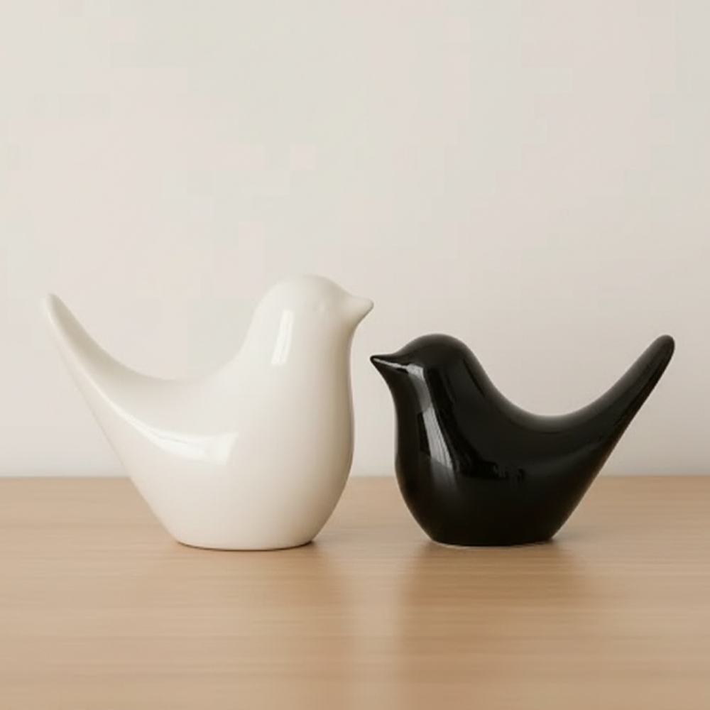 Two ceramic bird-shaped objects, one white and one black, on a wooden surface with a plain background.